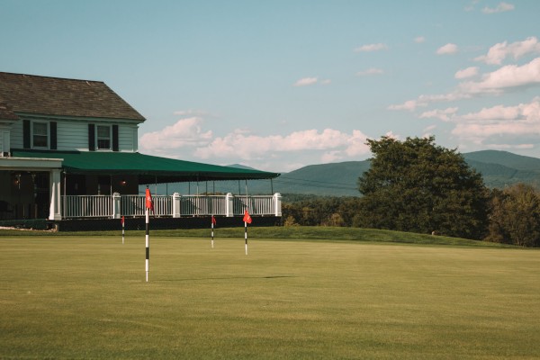A golf putting green with several holes and flags, next to a building, with mountains and trees in the background.